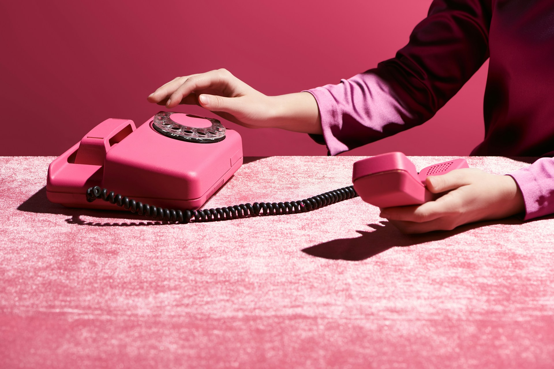 A pink retro rotary telephone on a pink textured surface, with a person's hands holding the receiver and touching the base, against a solid pink background.