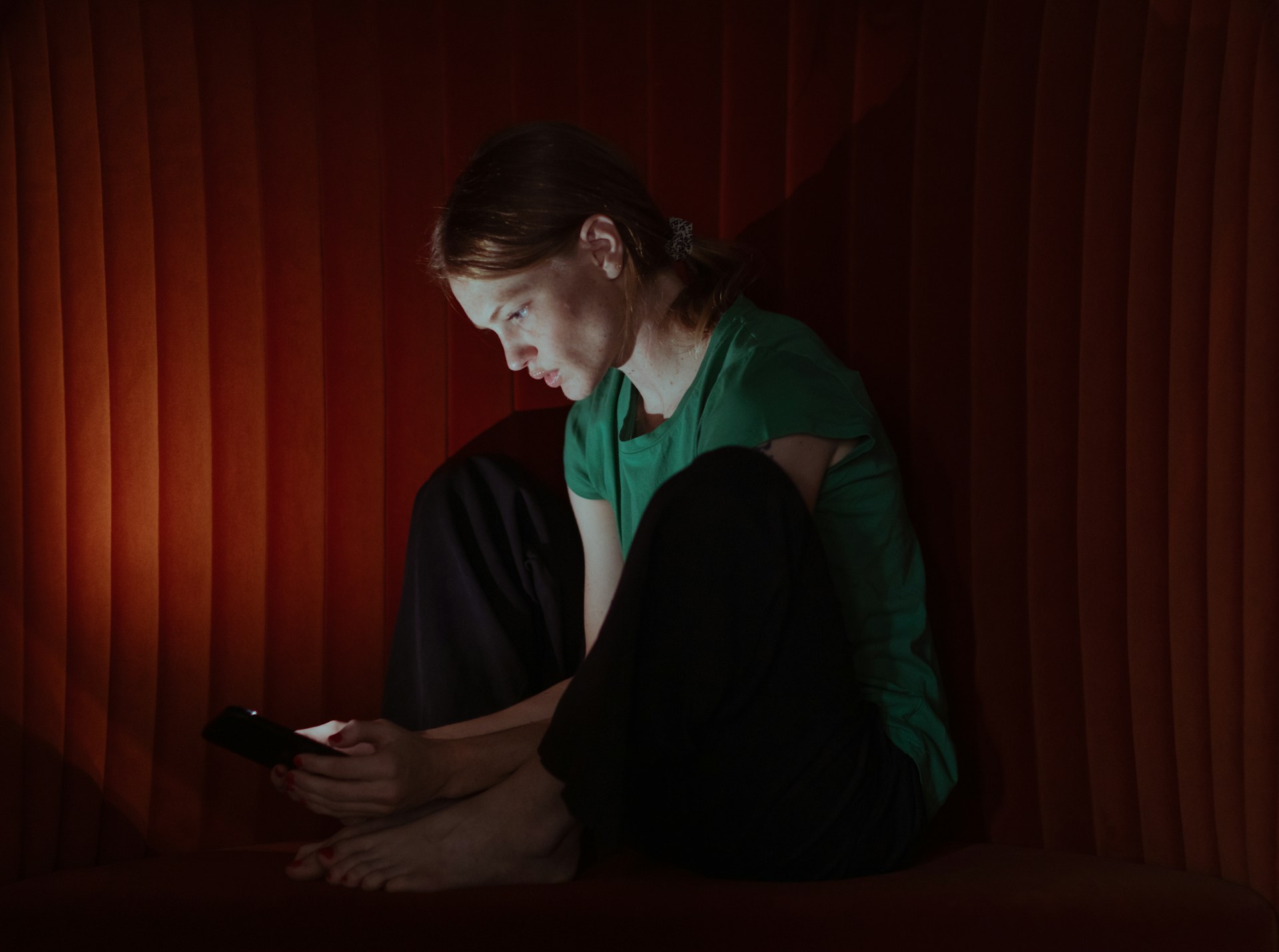 A woman sits barefoot against a dark red ribbed wall, looking down at a phone in her hands. She is wearing a green short-sleeve top and black pants.